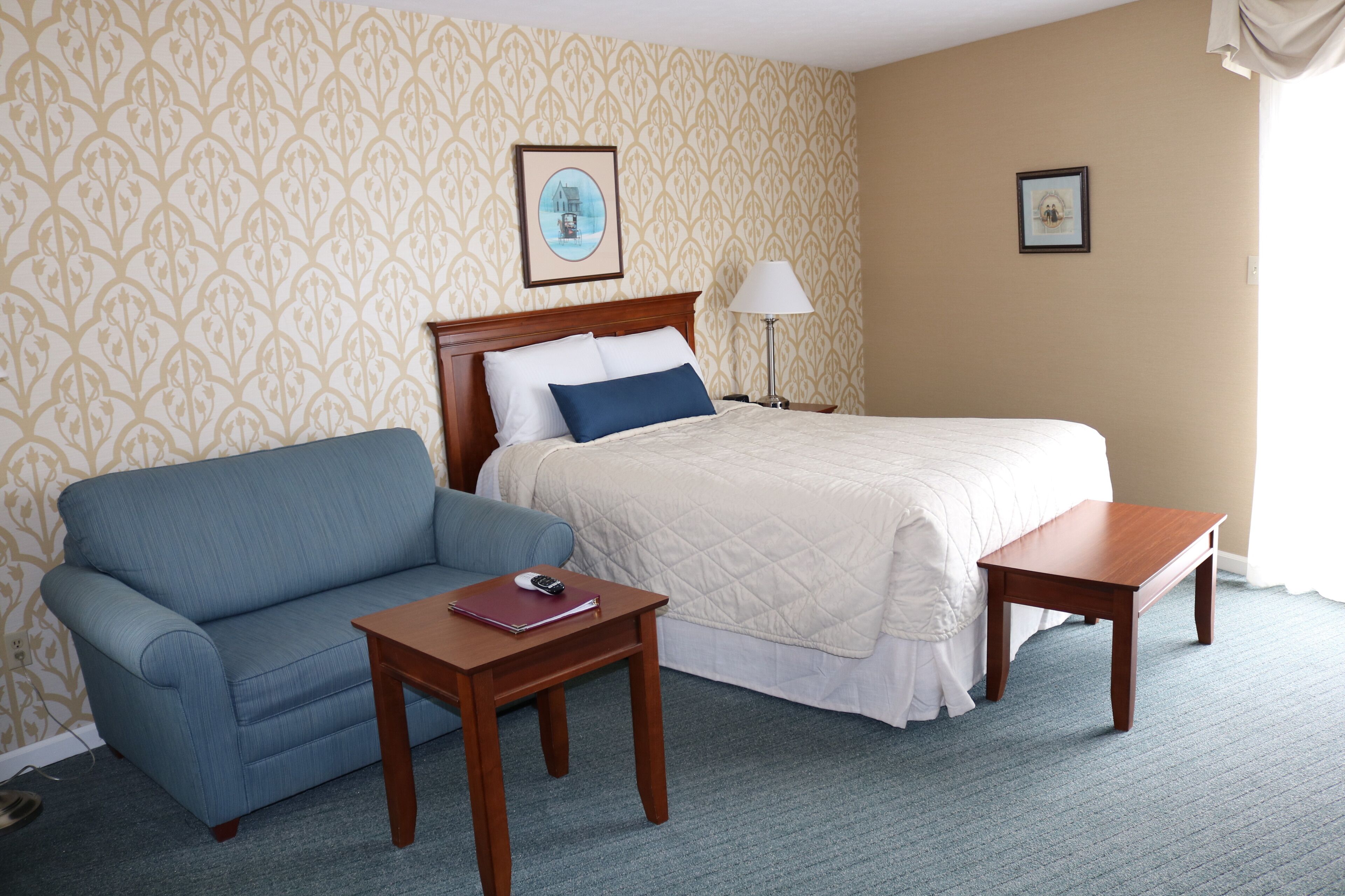 A hotel room with a bed, a blue sofa, a wooden nightstand, and a lamp.
