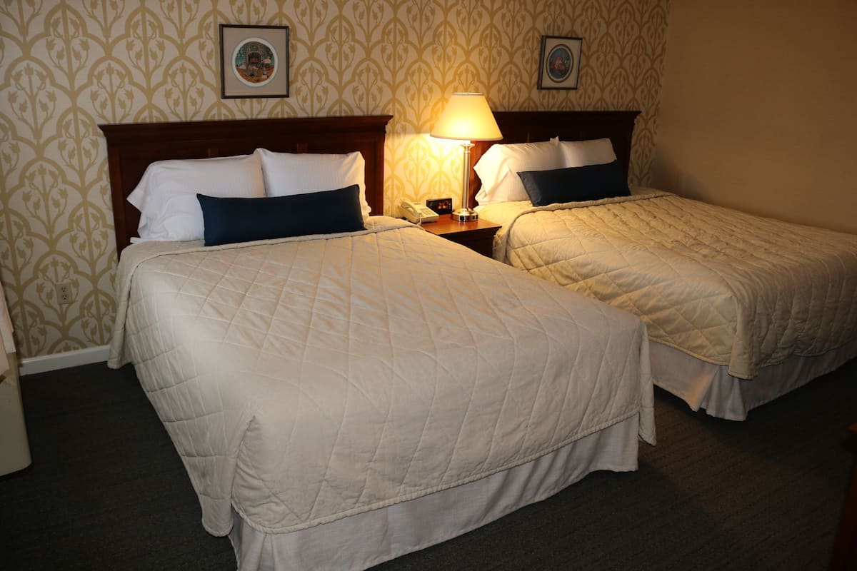 Two single beds with white and blue bedding, a nightstand with a lamp, and a patterned wallpaper.