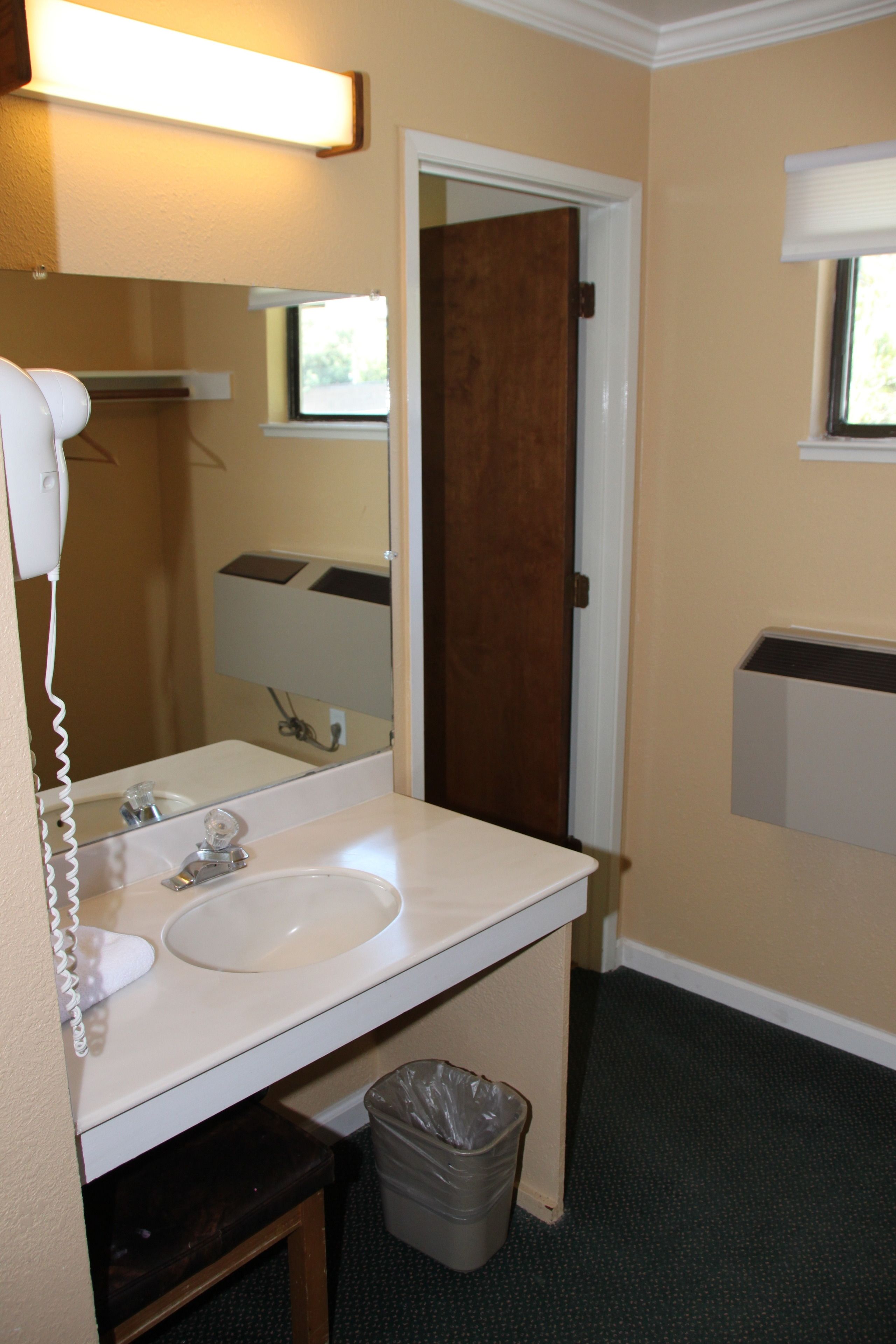 A bathroom with a white sink, a mirror, a hairdryer, a trash bin, and a window with blinds.