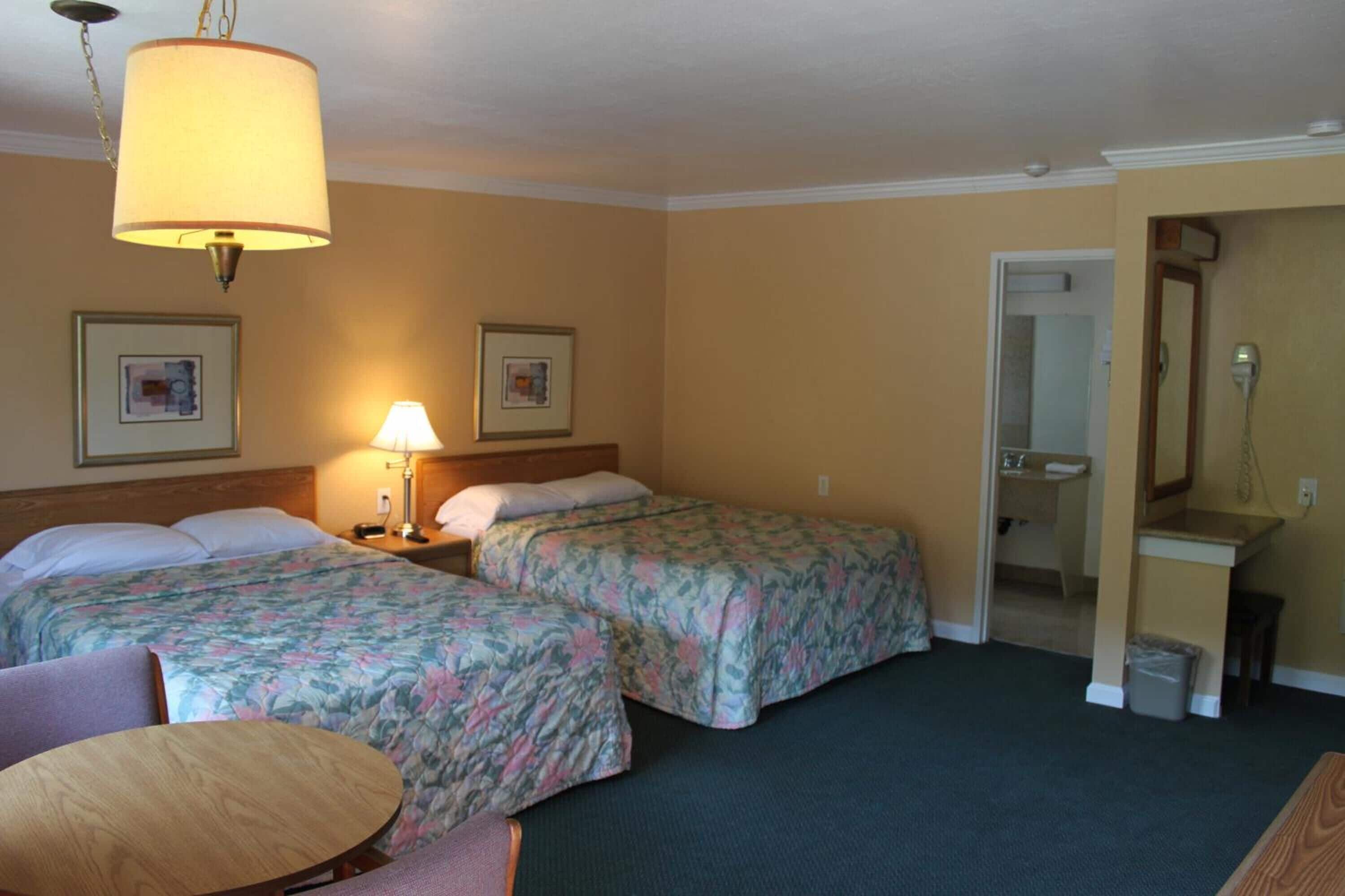 A hotel room with two beds, a round table, a lamp, and a bathroom visible through an open door.
