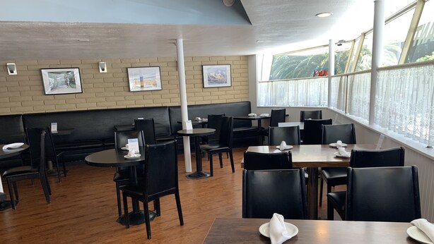 A restaurant with wooden floors, black chairs, and tables. There are framed pictures on the wall and a window with curtains.
