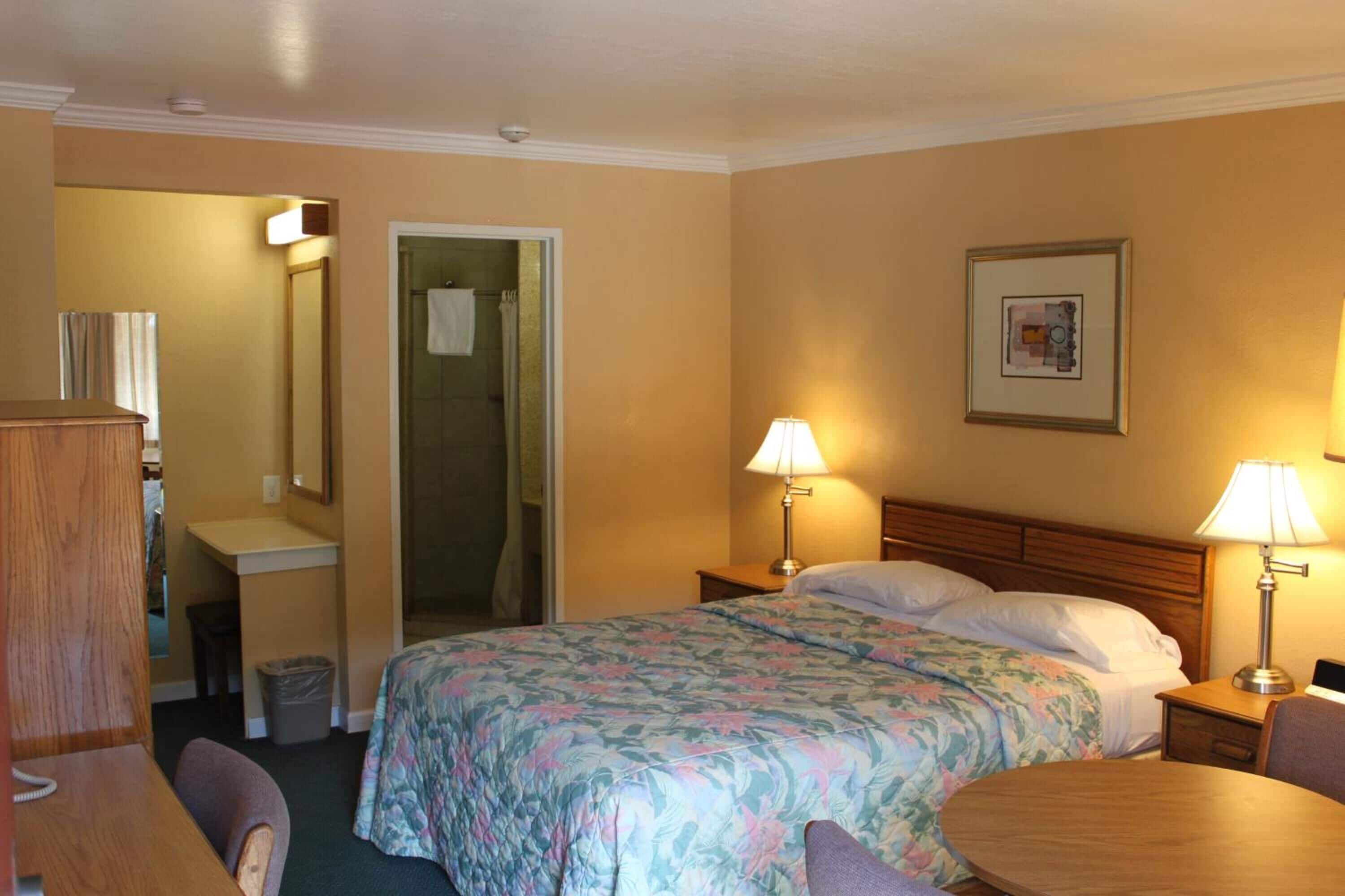 Habitación de hotel con cama, lámparas de noche, escritorio, silla, baño y un cuadro en la pared.