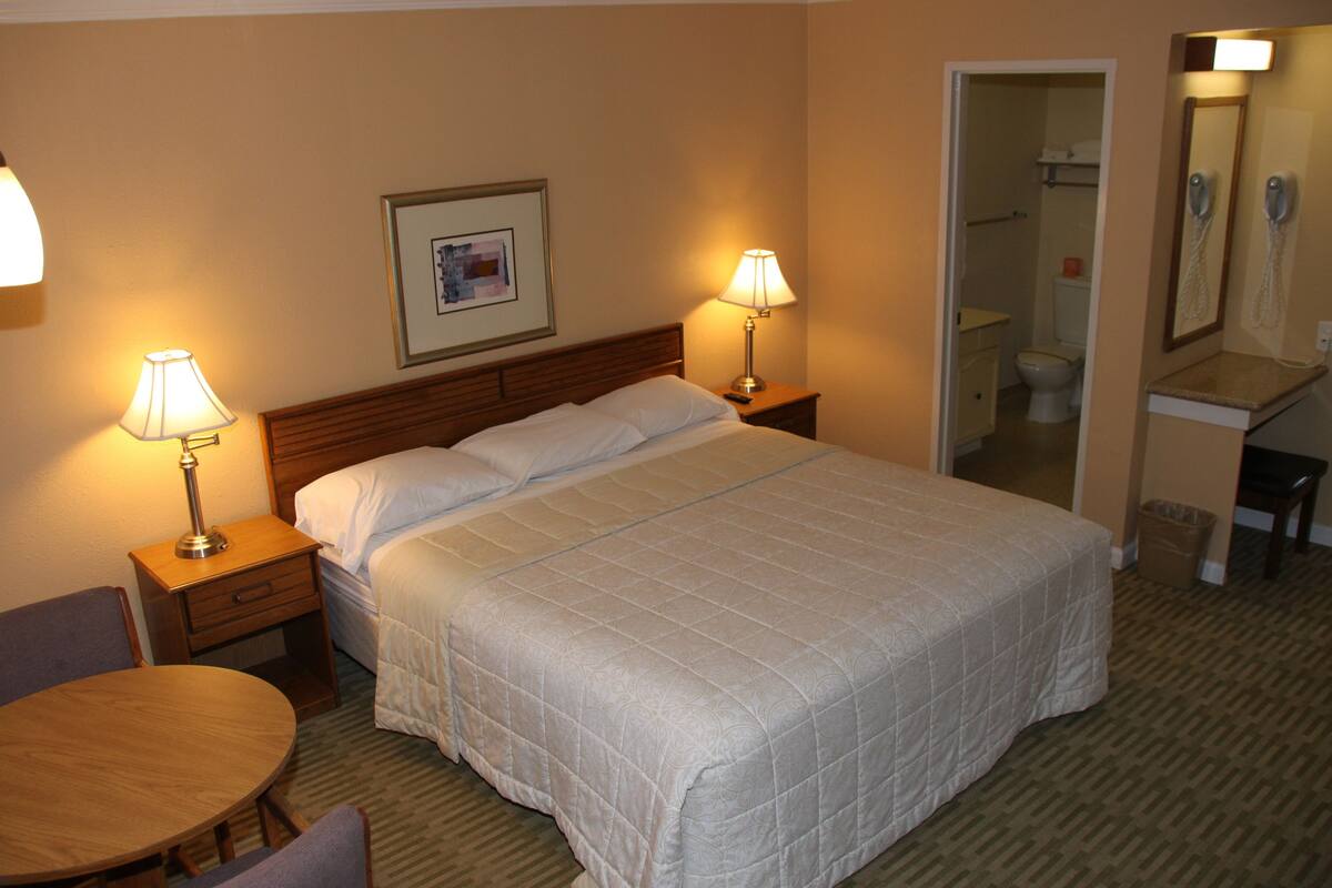 A hotel room with a large bed, two bedside tables with lamps, a small round table, and a chair. There is a framed picture on the wall above the bed.
