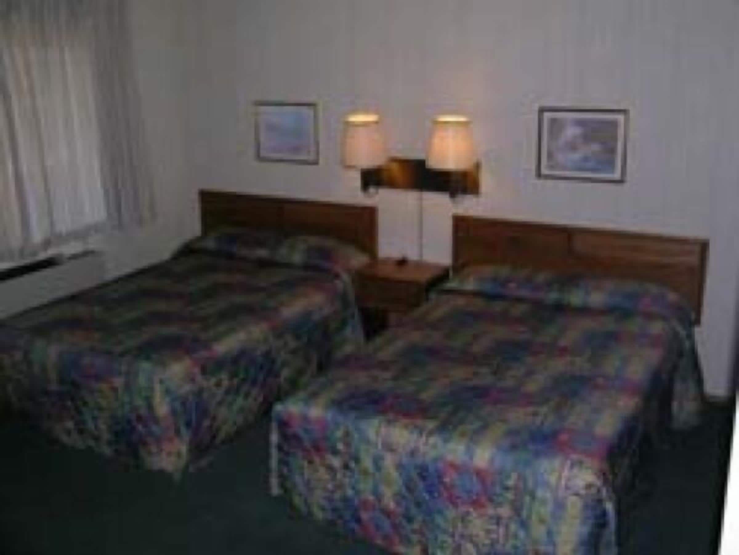 A hotel room with two single beds, a wooden headboard, a nightstand with two lamps, and framed pictures on the wall.