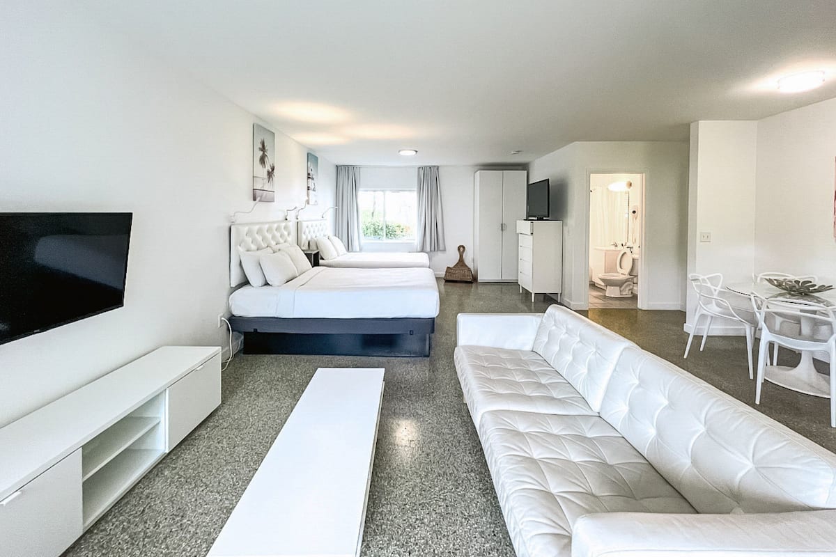 Habitación de hotel moderna con dos camas, televisor, sofá, mesa de comedor y baño visible a través de una puerta abierta.