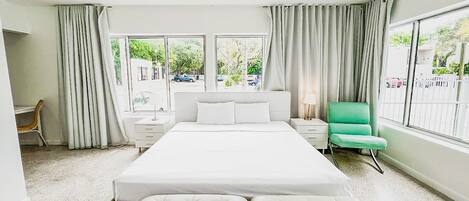 A modern hotel room with a large bed, a green armchair, a desk, and a view of the street through large windows.