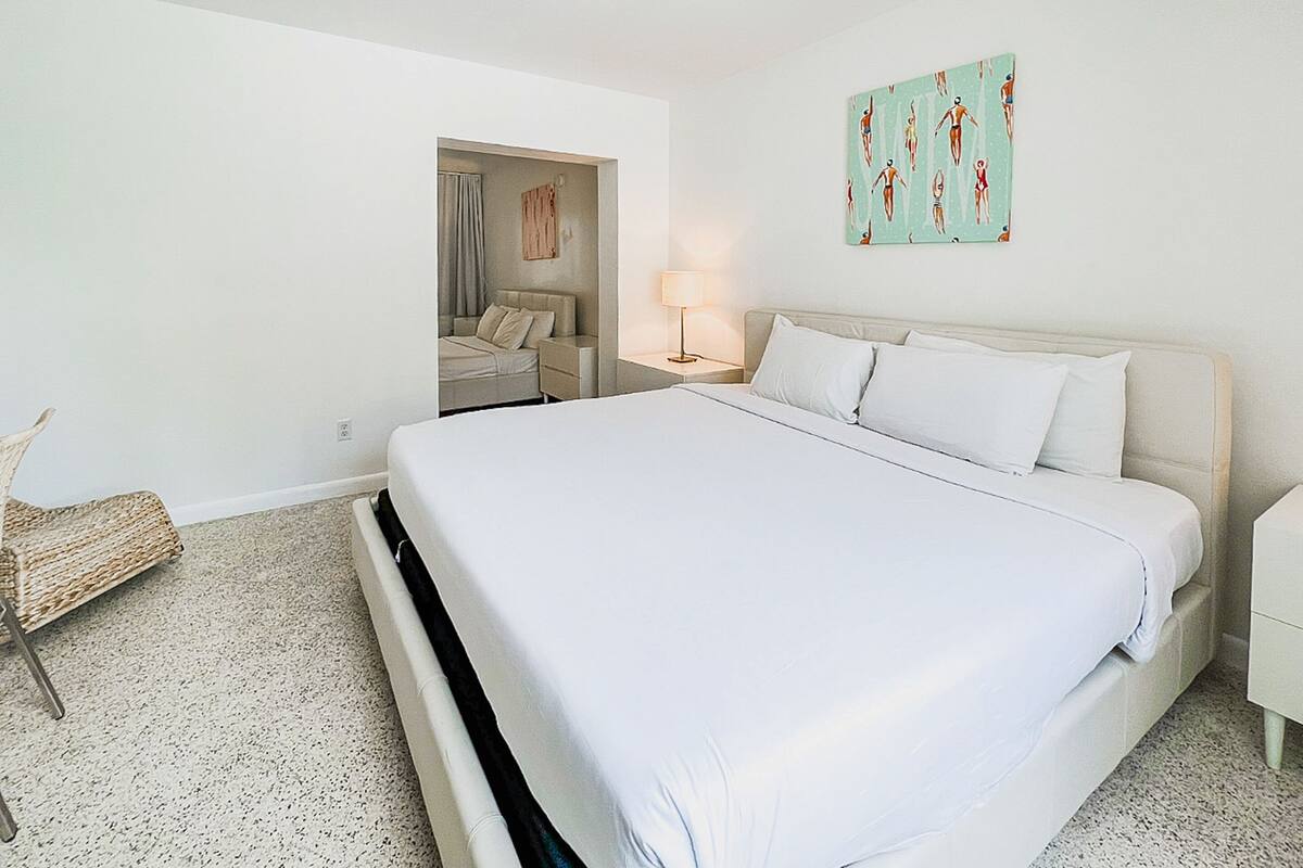 Habitación de hotel con una cama grande, una mesita de noche con lámpara, una silla y un cuadro en la pared.