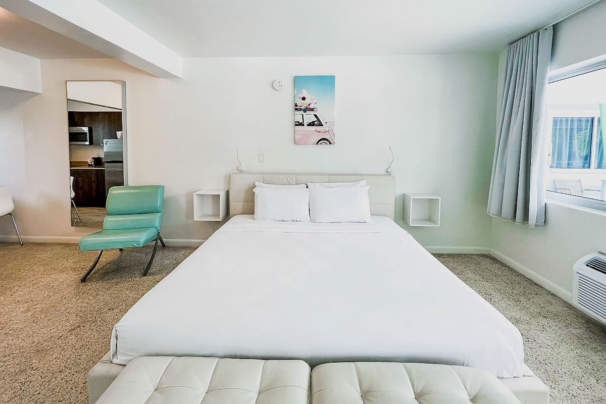 Una habitación de hotel con una cama grande, una silla, una mesita, un cuadro y una ventana con cortinas.
