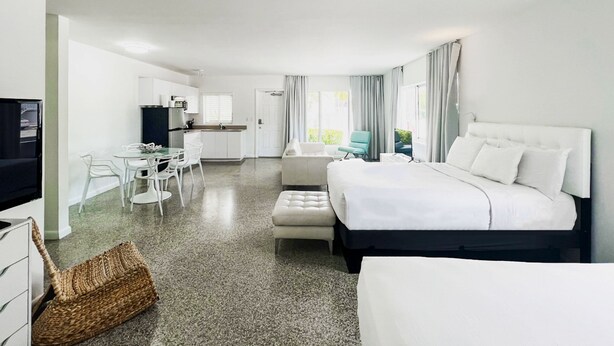 Habitación de hotel moderna con una cama grande, una zona de estar separada con mesa y sillas, una kitchenette y vistas al exterior.