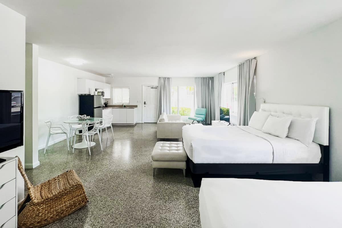 Habitación de hotel moderna con una cama grande, una zona de estar separada con mesa y sillas, una kitchenette y vistas al exterior.