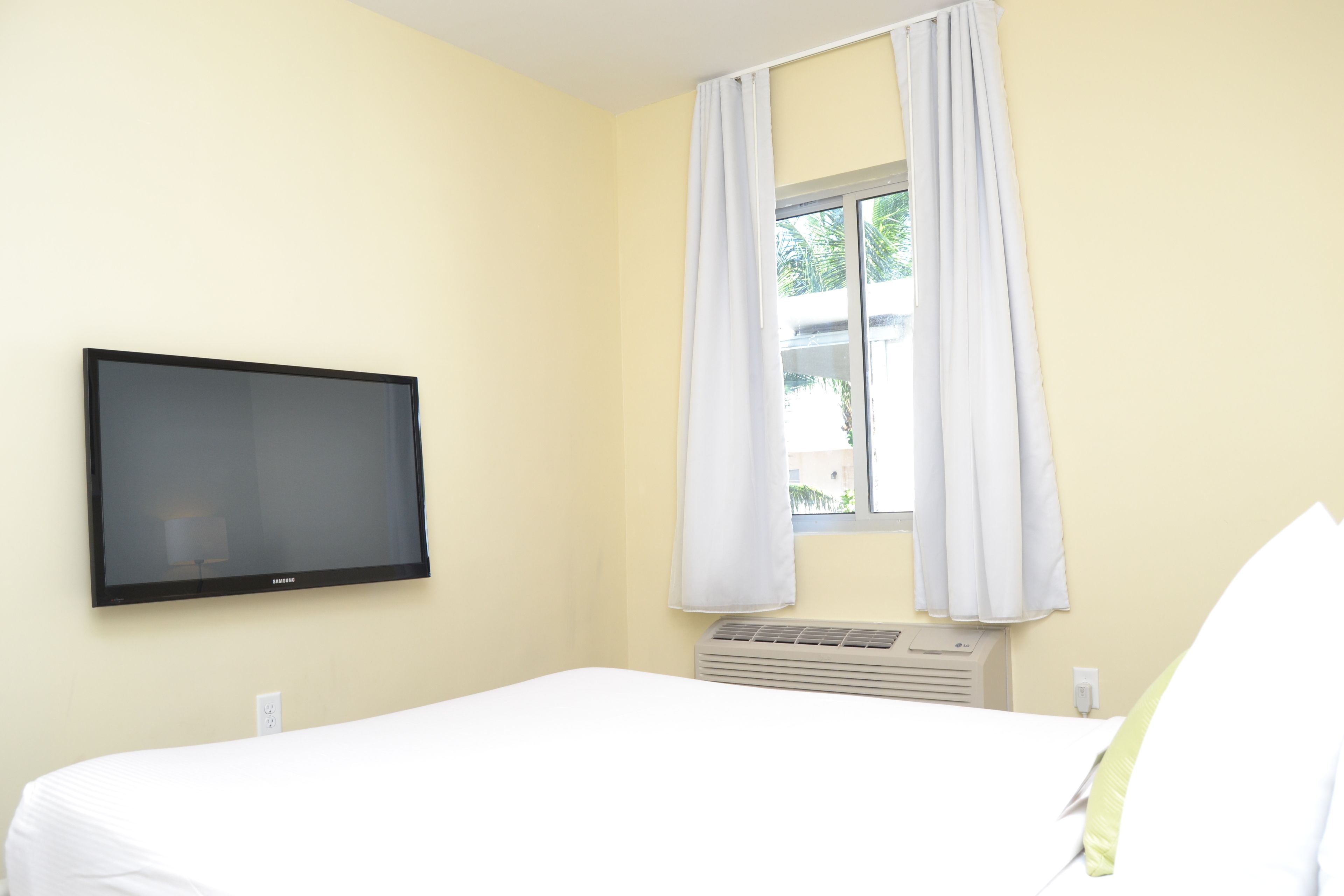 A hotel room with a bed, a flat-screen TV mounted on the wall, a window with white curtains, and a wall-mounted air conditioning unit.