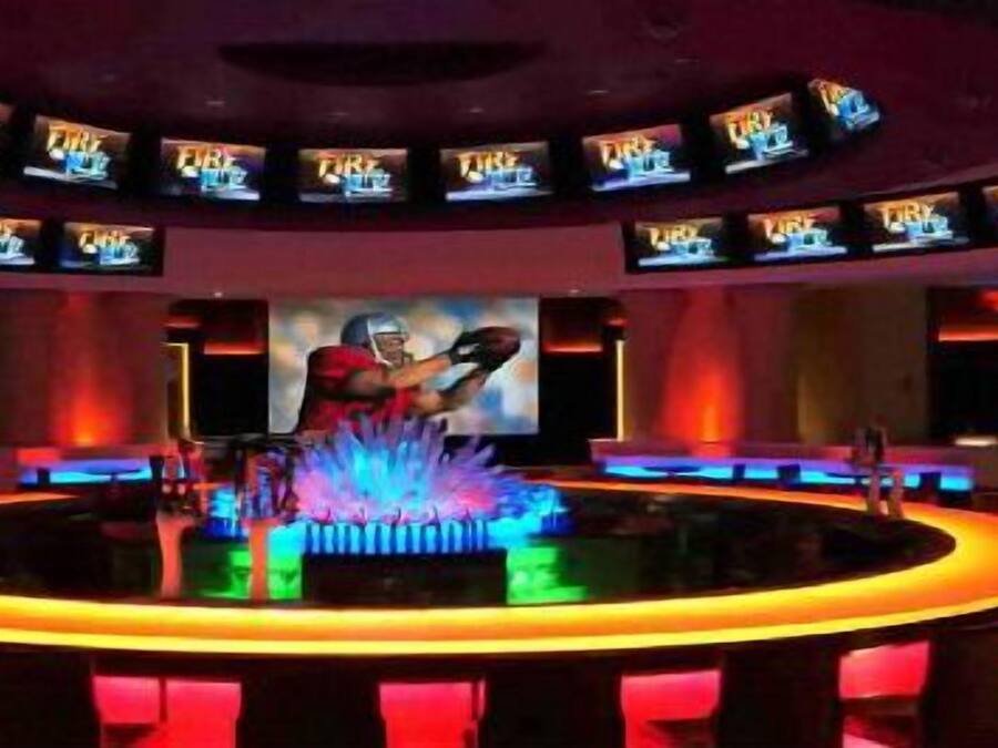 A bar area with multiple screens displaying content, a central bar counter, and a person in a red uniform.
