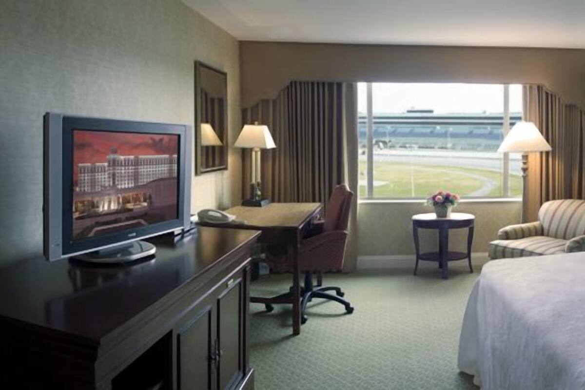 Habitación de hotel con cama, escritorio con televisor, silla, mesita con lámpara y ventana con vista al aeropuerto.