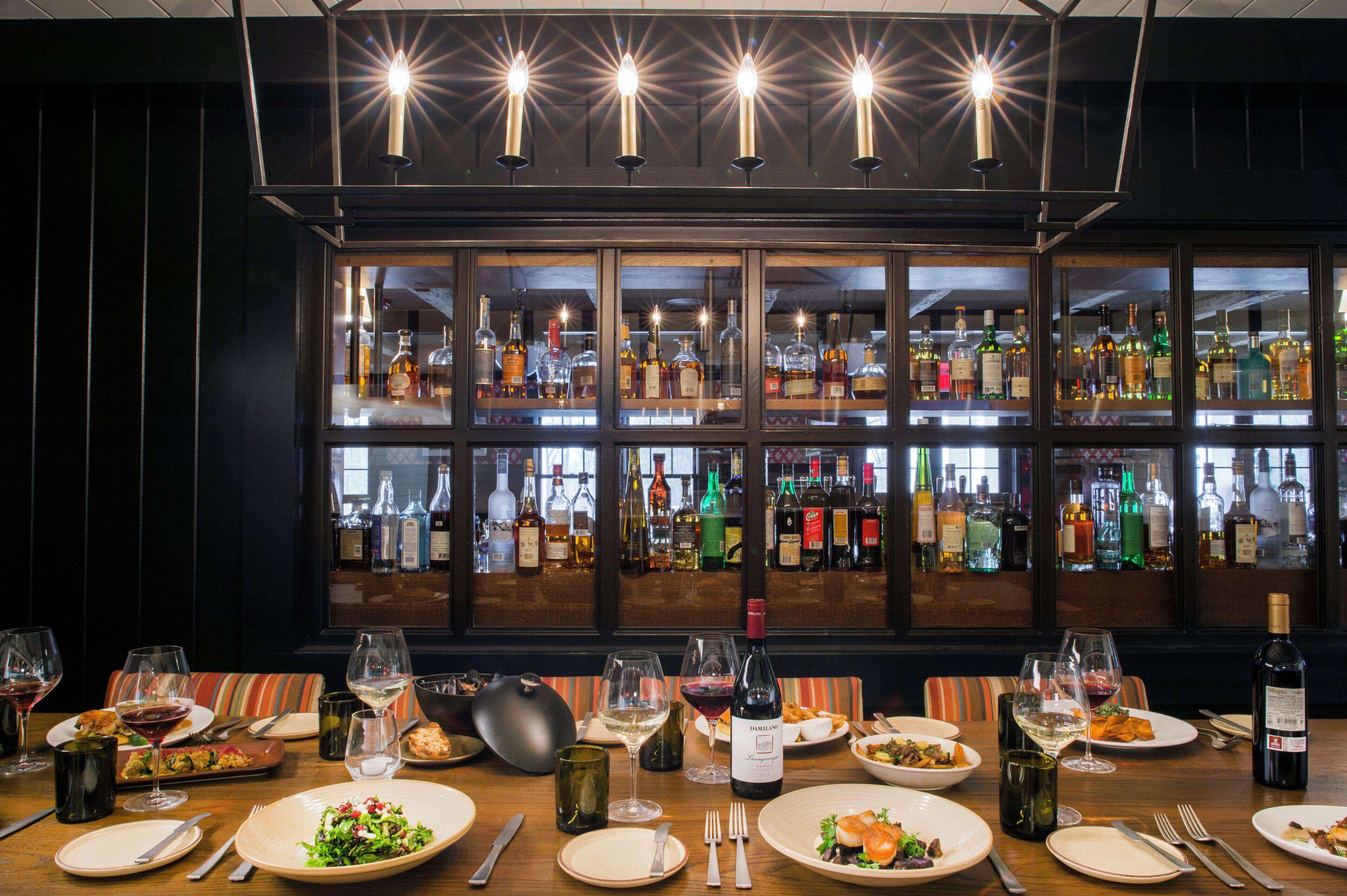 A restaurant interior with a bar area, multiple dining tables set with plates, glasses, and cutlery, and a large window displaying various bottles.