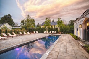 Una piscina con sillones de descanso y sombrillas, rodeada de árboles y un edificio.