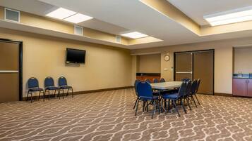 A conference room with a patterned carpet, a flat-screen TV mounted on the wall, a clock, and a kitchenette area.