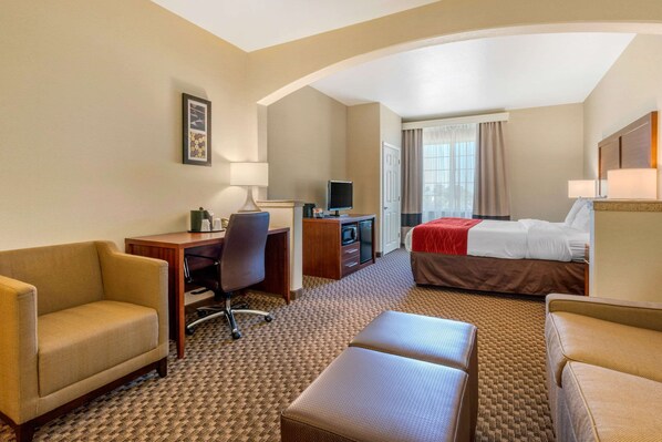 Habitación de hotel con sofá, escritorio con computadora, televisor, cama con una colcha roja y ventana con cortinas.