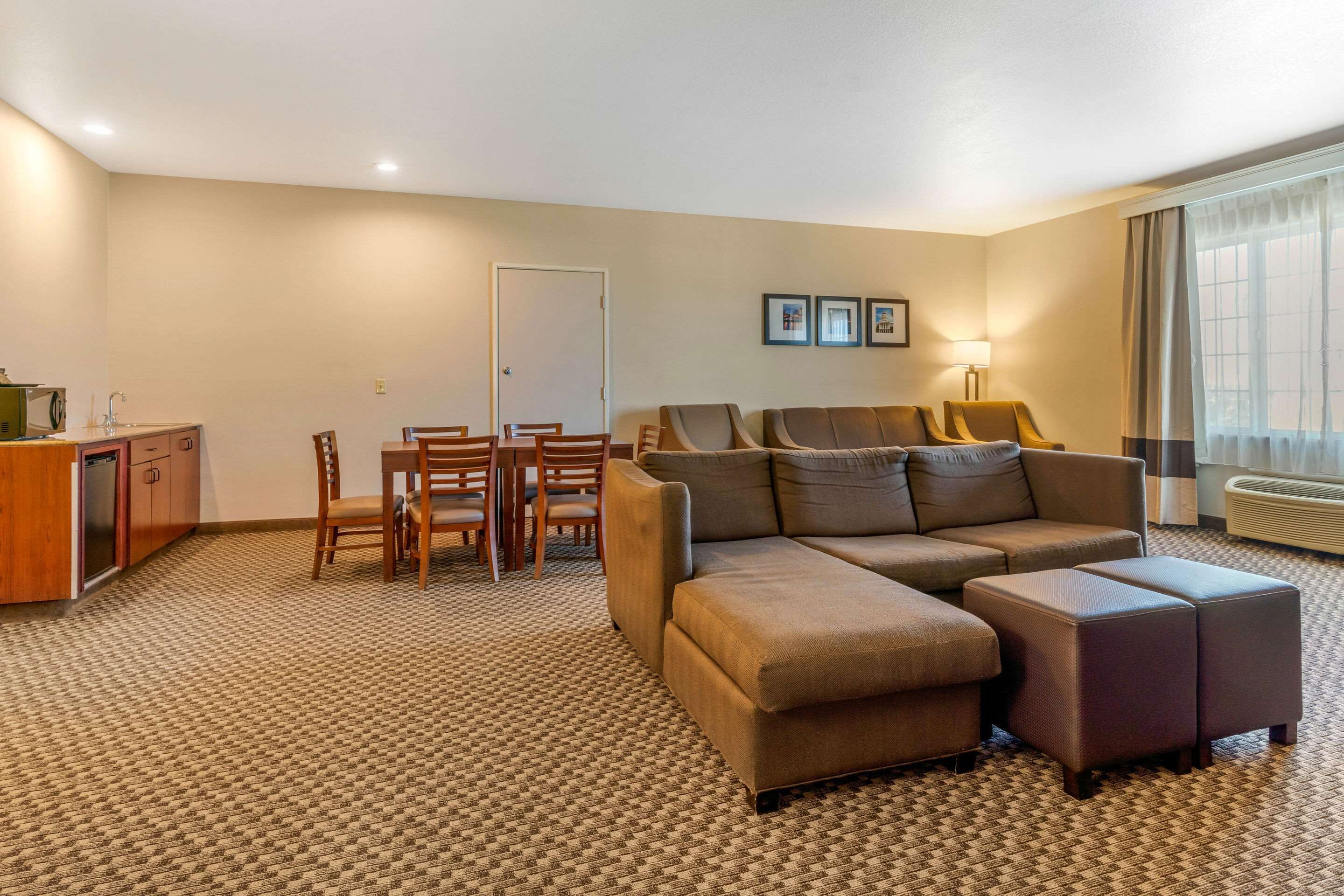 Habitación de hotel con sofá, reposapiés, mesa de comedor, sillas, kitchenette y un gran ventanal con cortinas.