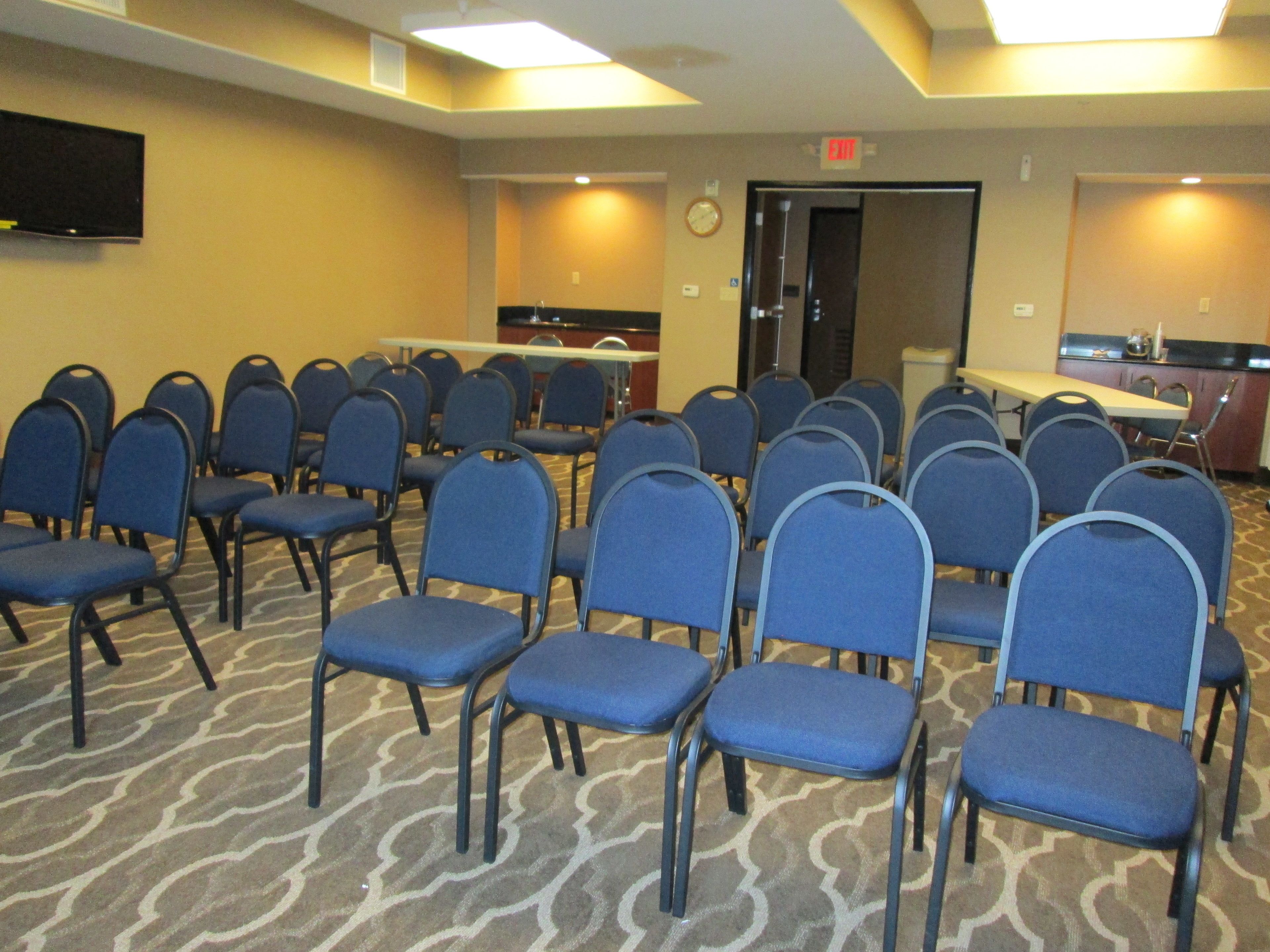 Un salón de conferencias con filas de sillas azules, una mesa, un reloj y un televisor en la pared.