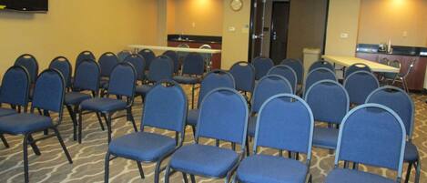 Un salón de conferencias con filas de sillas azules, una mesa, un reloj y un televisor en la pared.