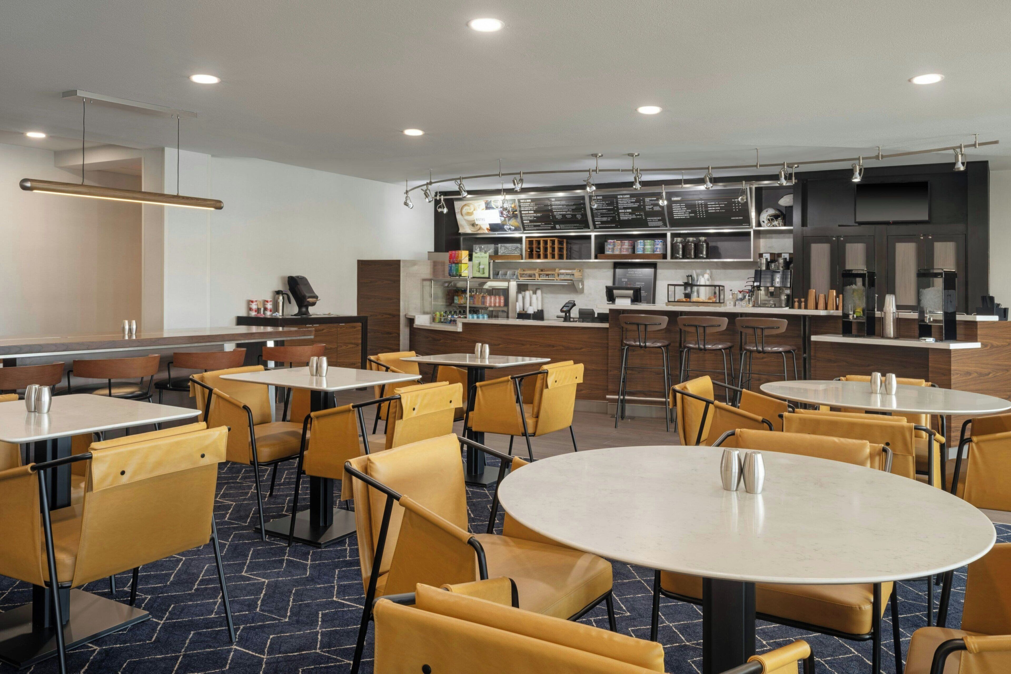 Un restaurante moderno con mesas redondas y sillas amarillas, una barra con menú y una zona de cocina.
