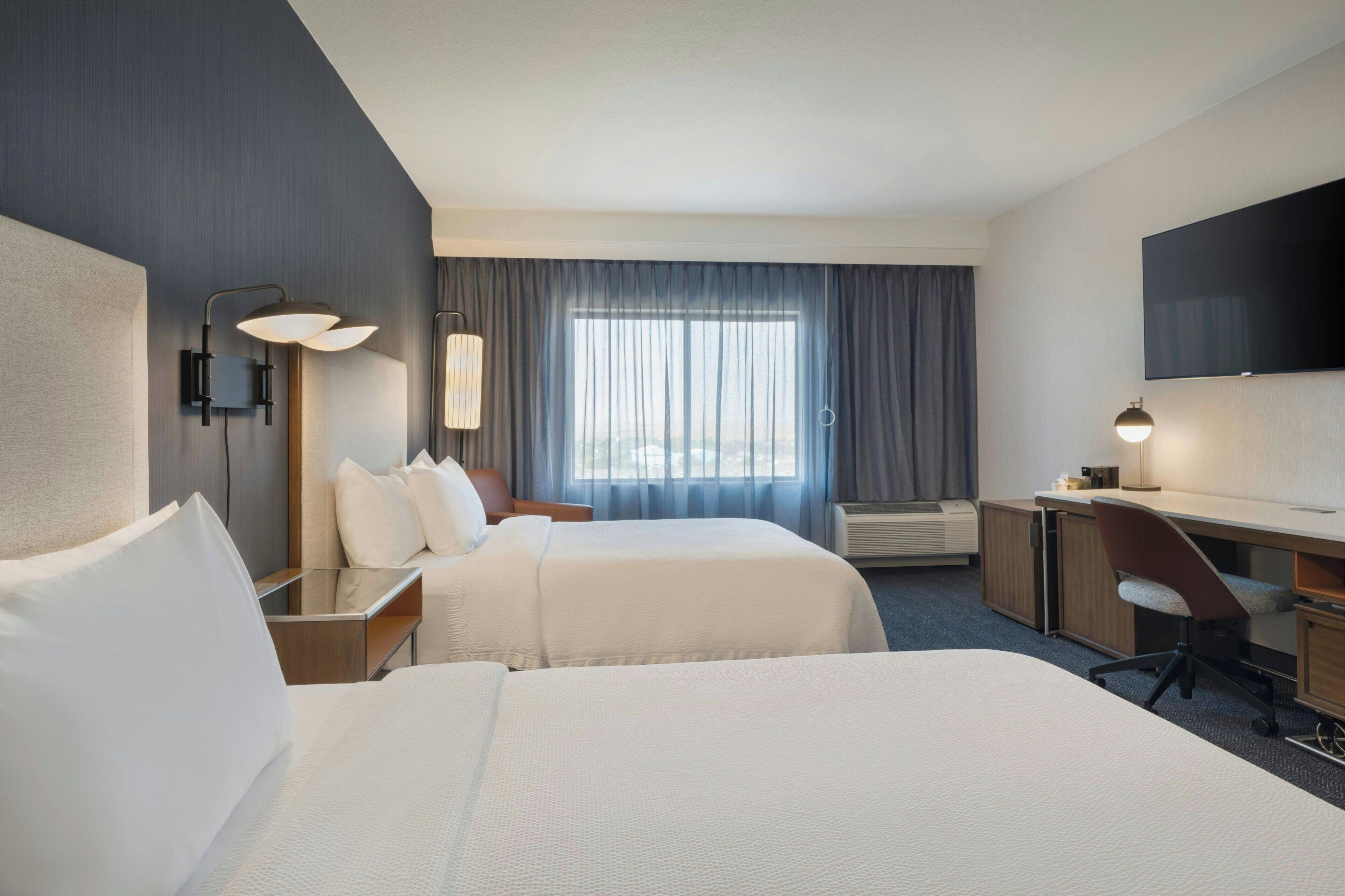 A hotel room with two beds, a desk, a chair, a flat-screen TV, and a large window with curtains.