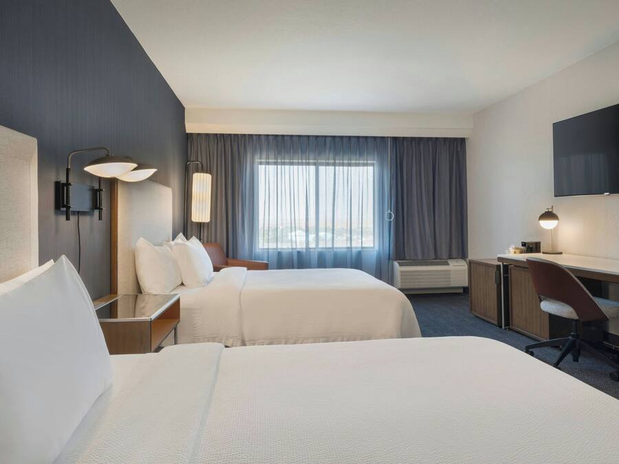 A hotel room with two beds, a desk, a chair, a flat-screen TV, and a large window with curtains.