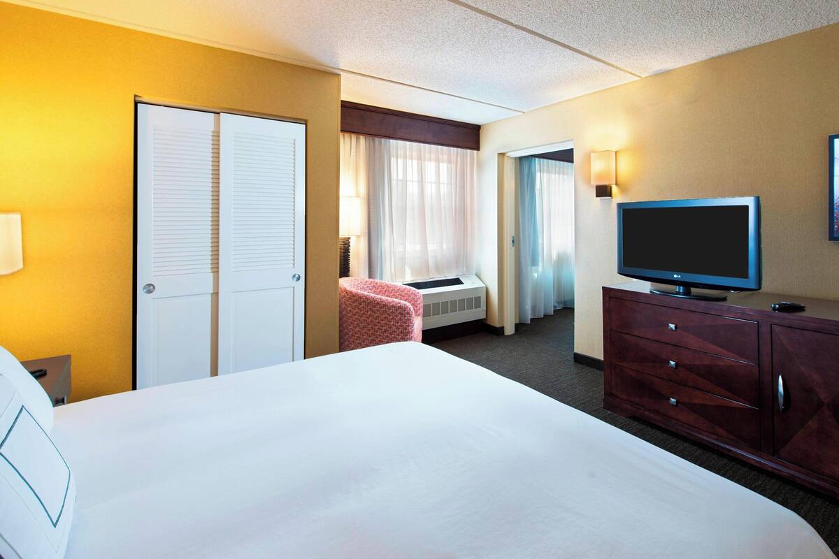 Habitación de hotel con una cama grande, un televisor en un aparador de madera, un escritorio con lámpara y una ventana con cortinas.
