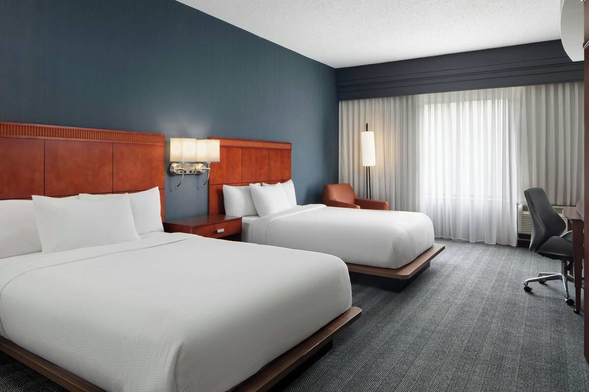 A hotel room with two beds, a desk, a chair, a lamp, and a large window with curtains.