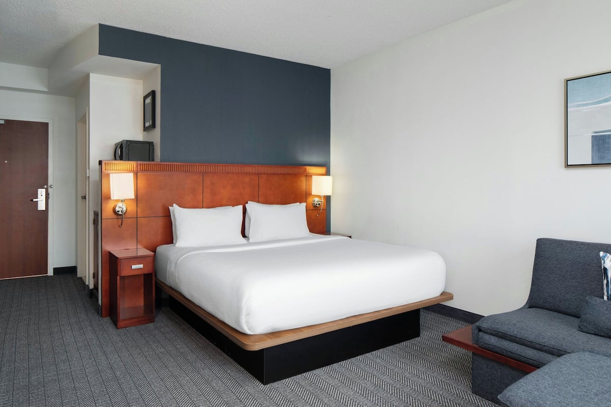 A hotel room with a large bed, two bedside lamps, a wooden headboard, a gray sofa, and a small table.