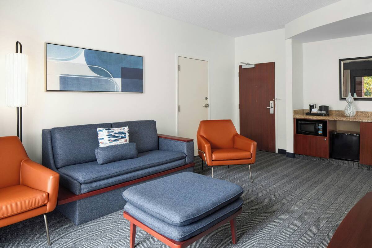A hotel room with a grey sofa, a blue ottoman, an orange armchair, a coffee machine, and a microwave.