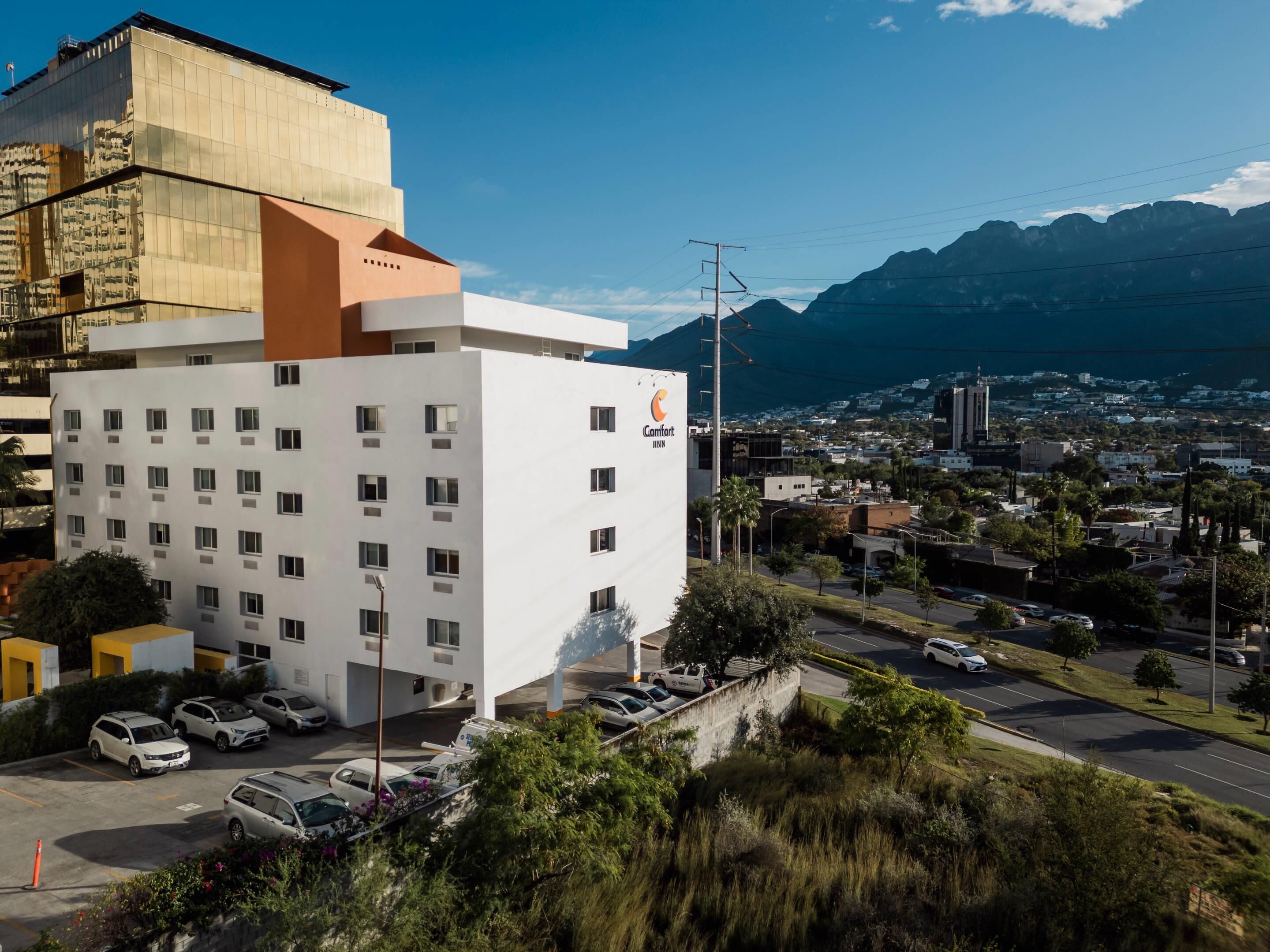 Un hotel moderno con fachada blanca y numerosas ventanas, ubicado en una ciudad con vistas a montañas y cielo despejado.