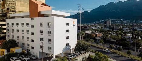 Un hotel moderno con fachada blanca y numerosas ventanas, ubicado en una ciudad con vistas a montañas y cielo despejado.