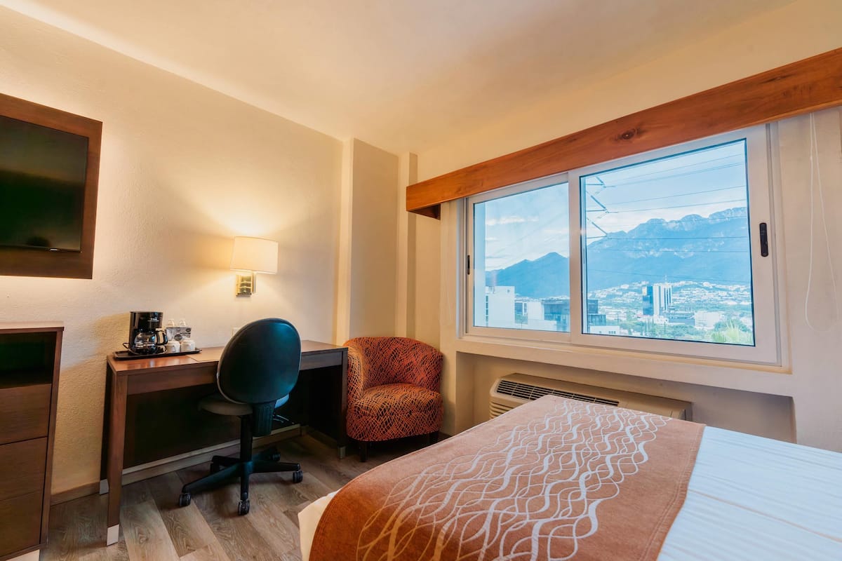 Habitación de hotel con cama, escritorio, silla y una ventana grande con vistas a la montaña.