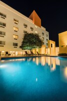 Zona de piscina nocturna con sillones reclinables y un edificio con múltiples ventanas y un árbol.