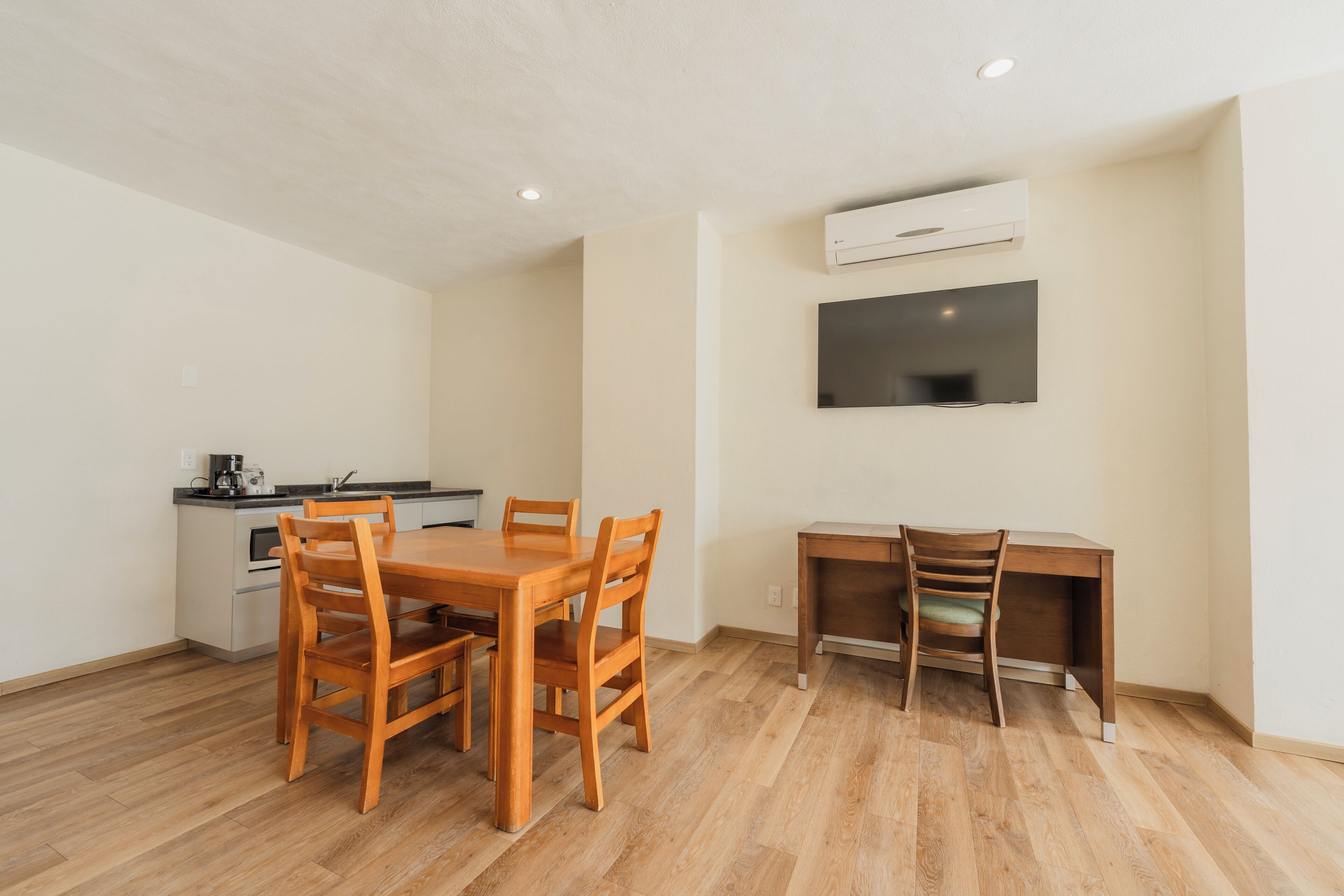 A compact living space with a wooden dining table, a kitchenette, a TV mounted on the wall, and a desk with a chair.