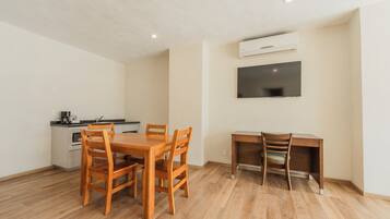 A compact living space with a wooden dining table, a kitchenette, a TV mounted on the wall, and a desk with a chair.