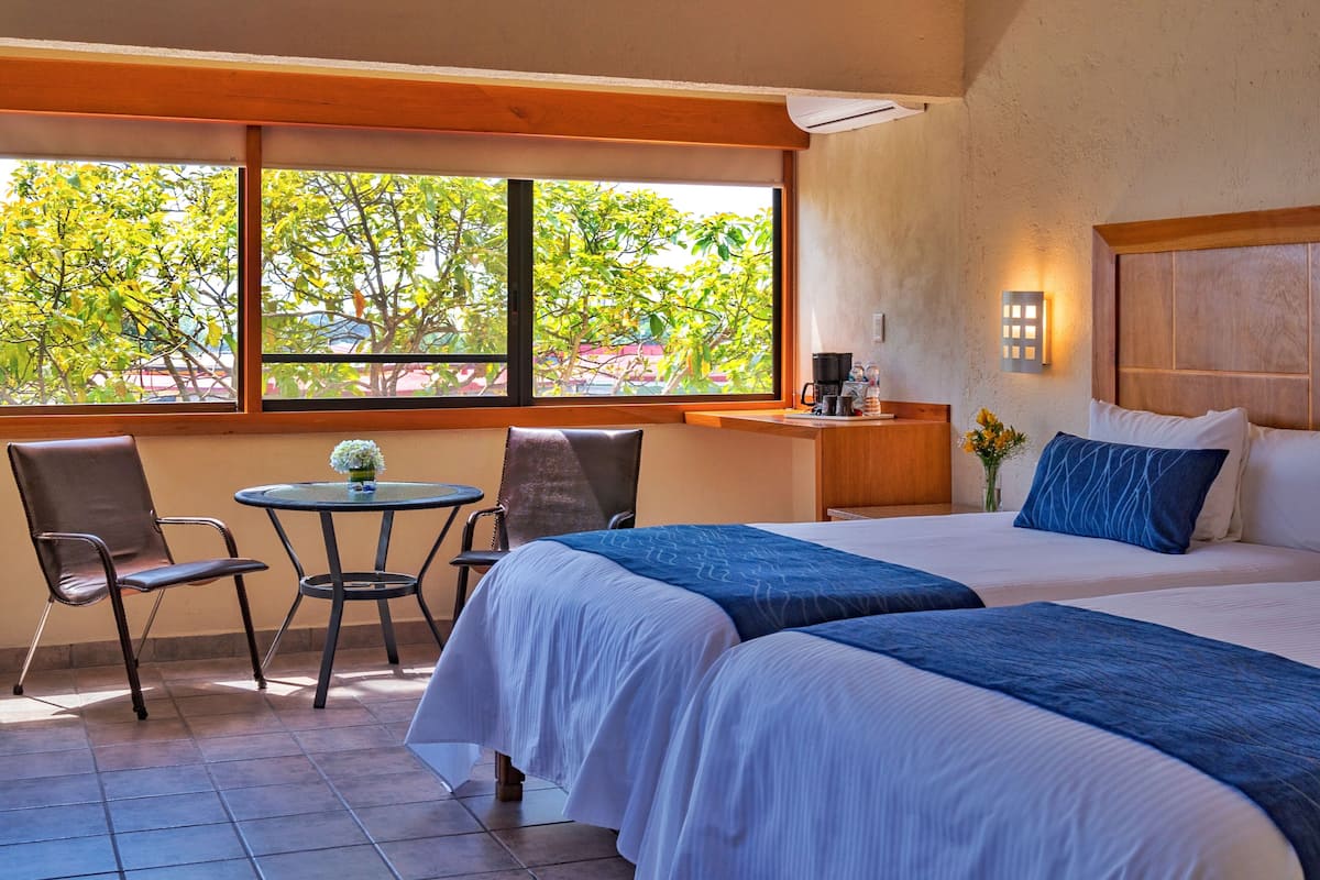 A hotel room with two beds, a small table, chairs, a window with a view of trees, and a wooden headboard.