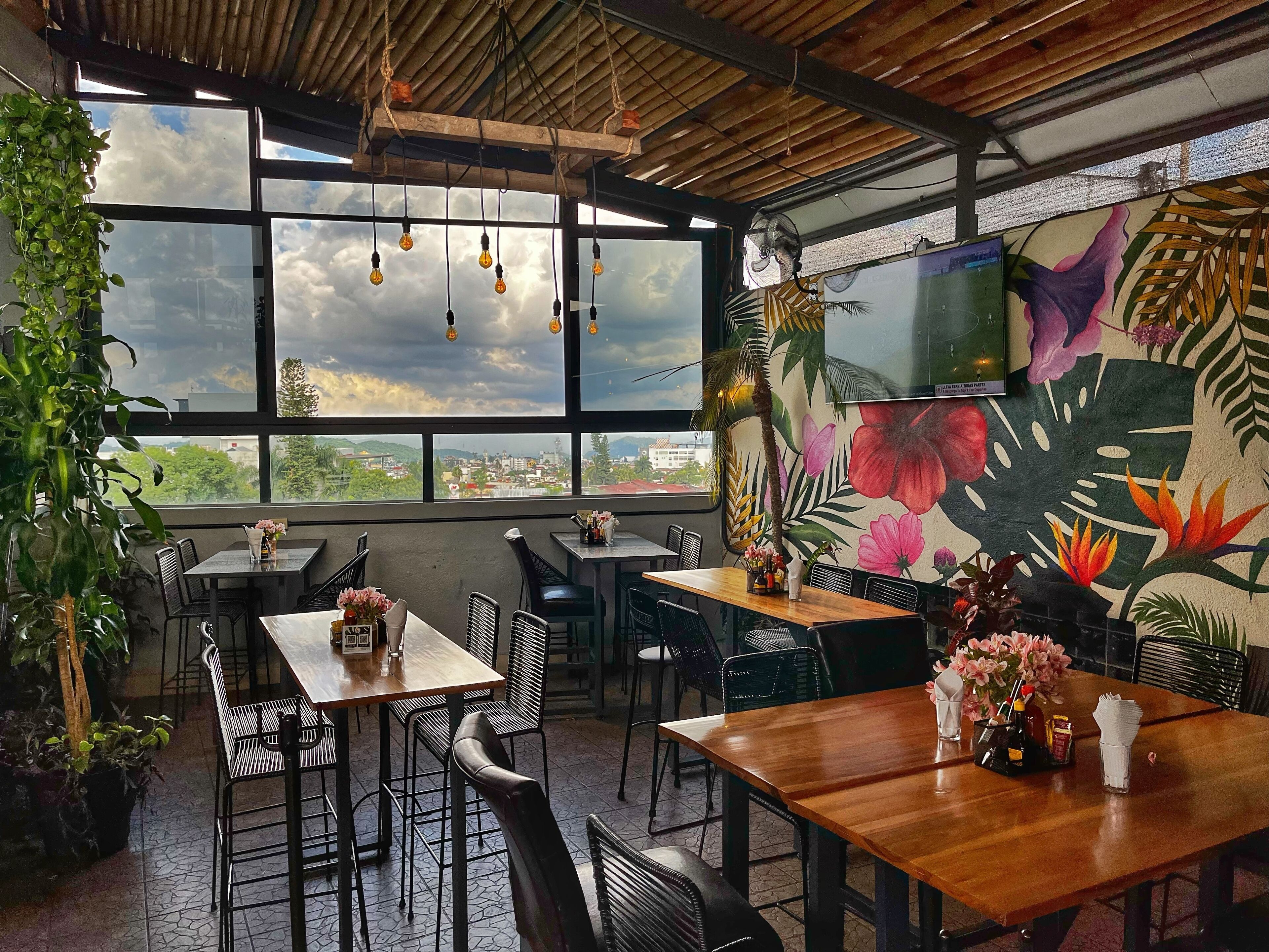 A restaurant with large windows, wooden tables, black chairs, and tropical-themed wall art.