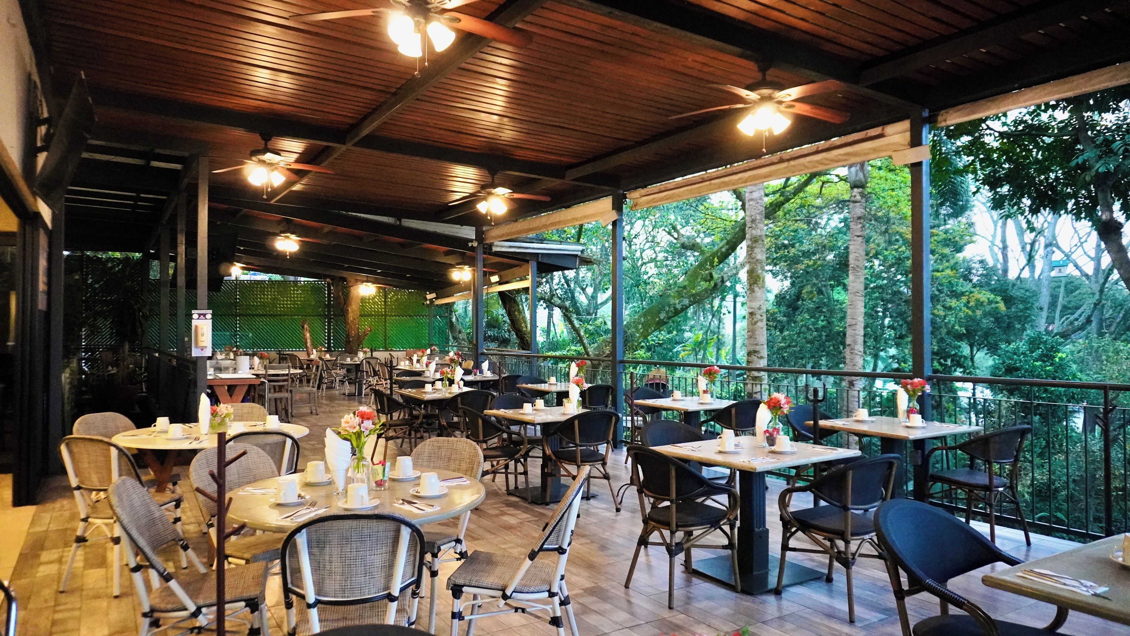An outdoor restaurant with wooden tables and chairs, large windows offering a view of the forest, and ceiling fans.