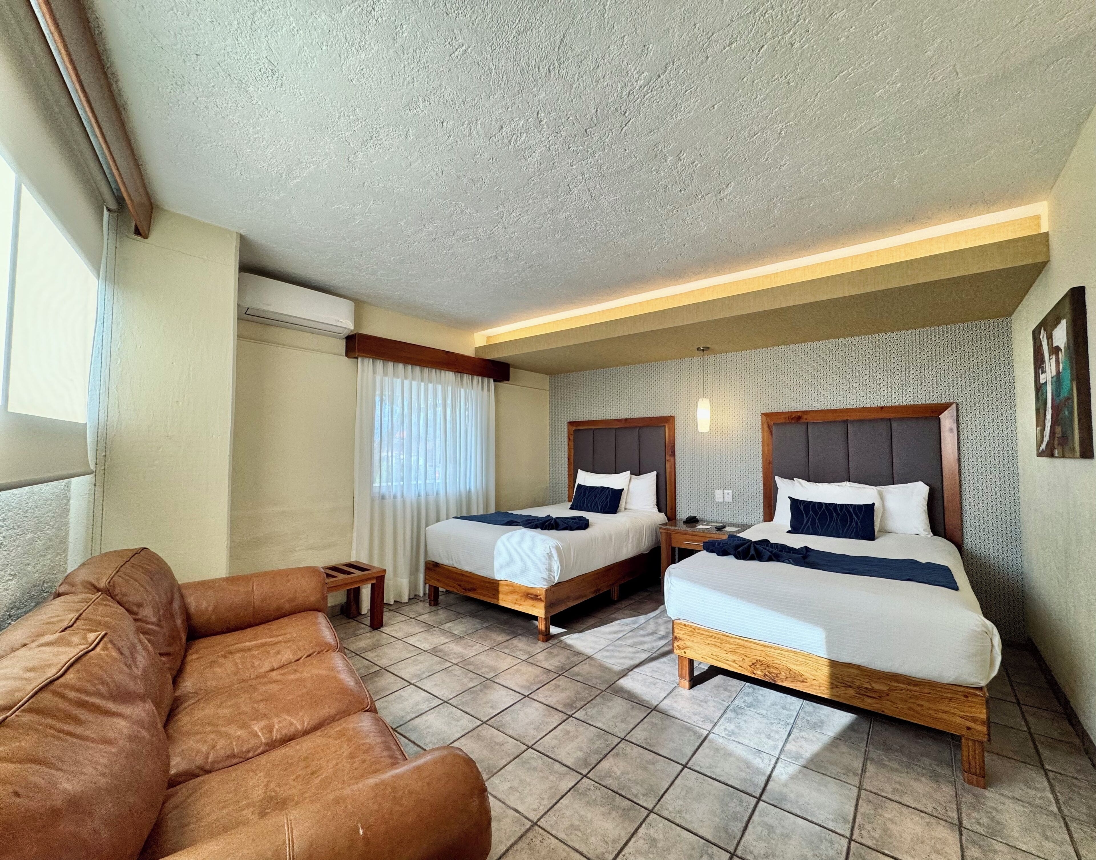 A hotel room with two beds, a brown leather sofa, a small table, and a window with curtains.