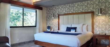 A hotel room with a large bed, a wooden headboard, a nightstand, a desk, a chair, and a window with curtains.