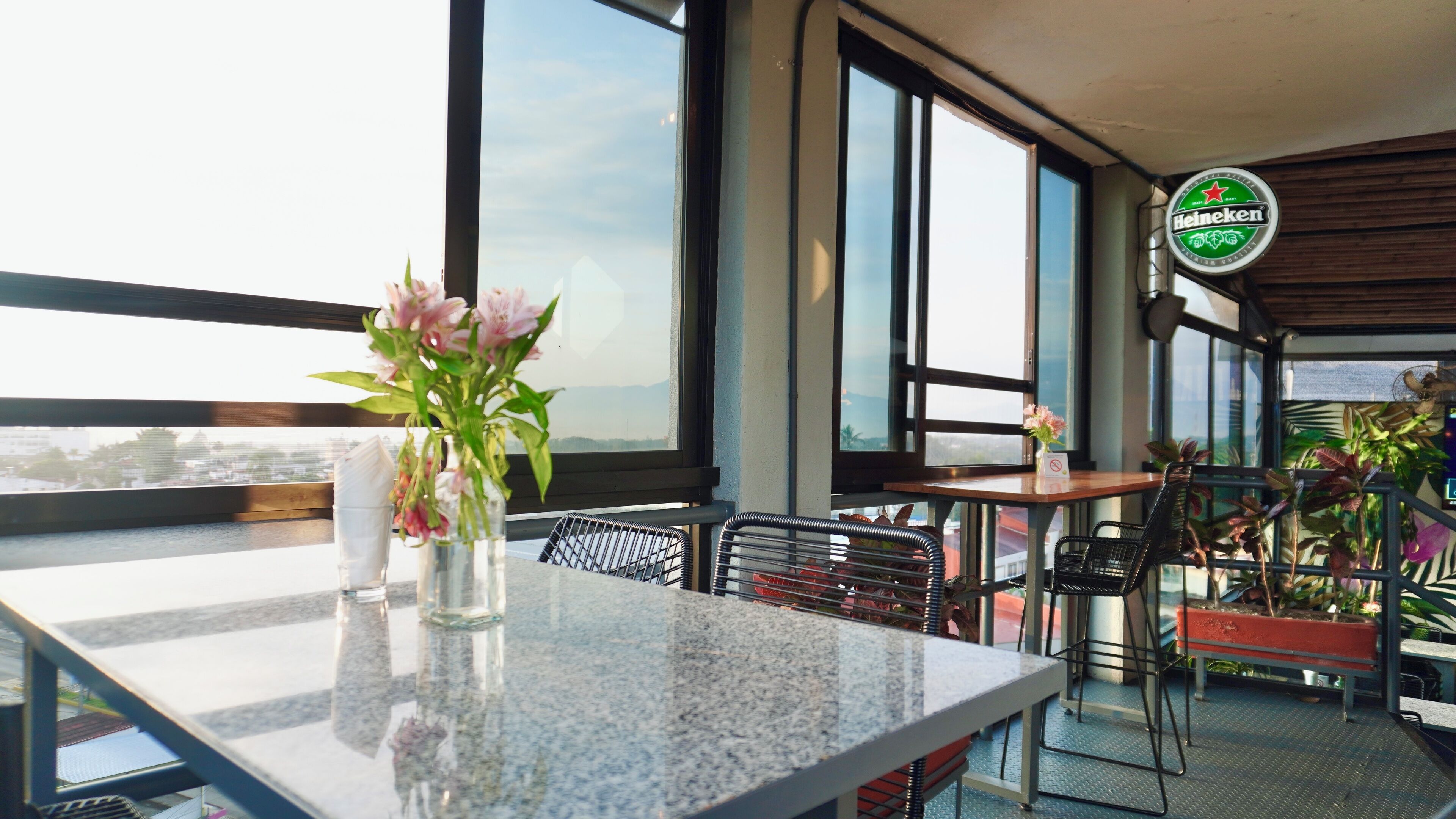 Un restaurante con vistas a la ciudad, un letrero de Heineken y una mesa con un jarrón de flores.