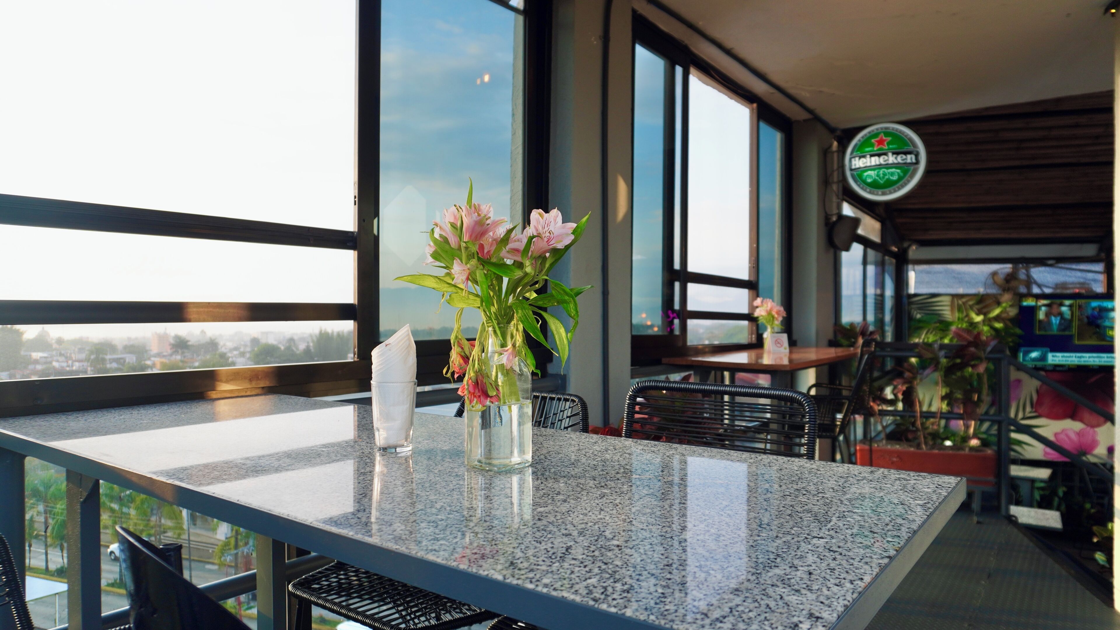 A restaurant with a view of the city, a Heineken sign, and a table with a vase of flowers.