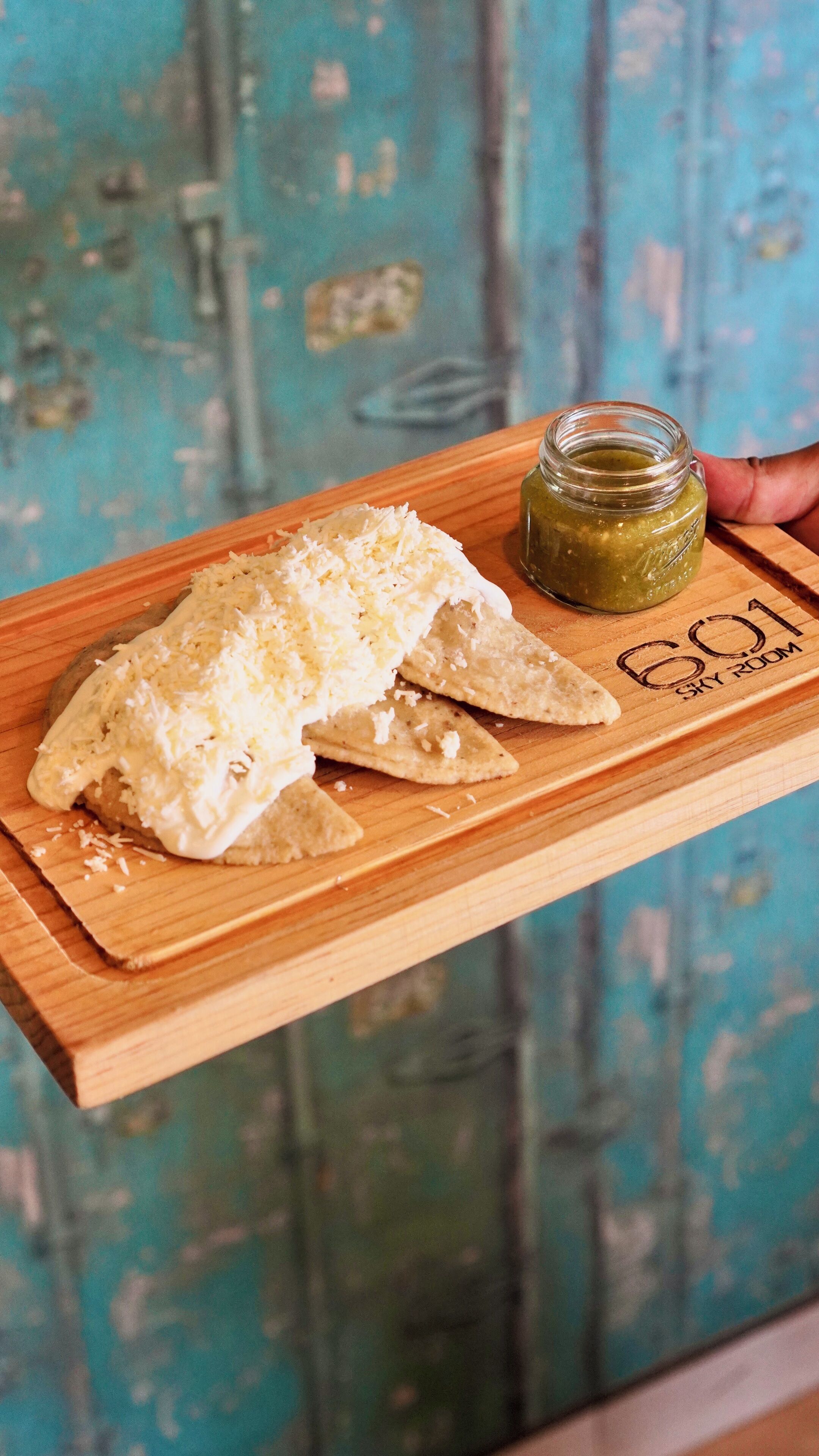 Una tabla de madera con un plato de nachos cubiertos de queso y un frasco pequeño de salsa verde.