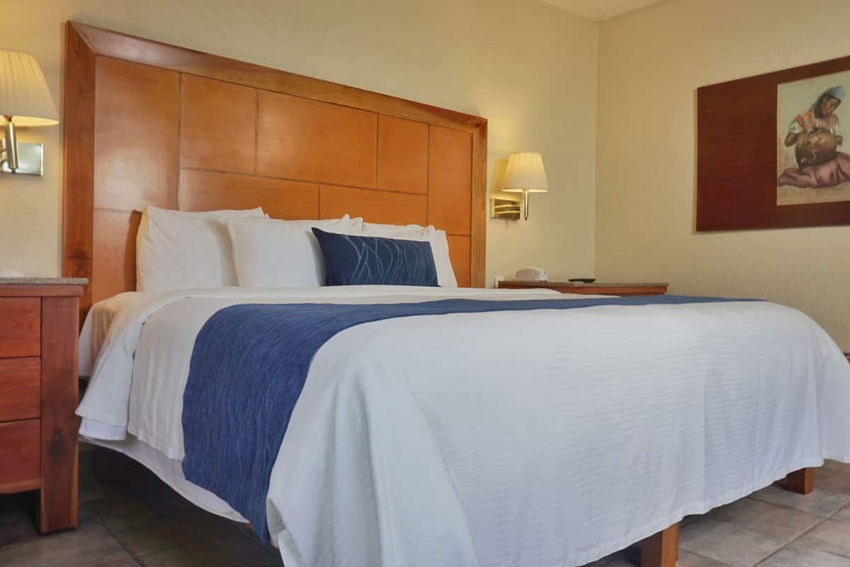 A hotel room with a large bed, wooden headboard, bedside lamps, a nightstand, a small table, and a painting on the wall.