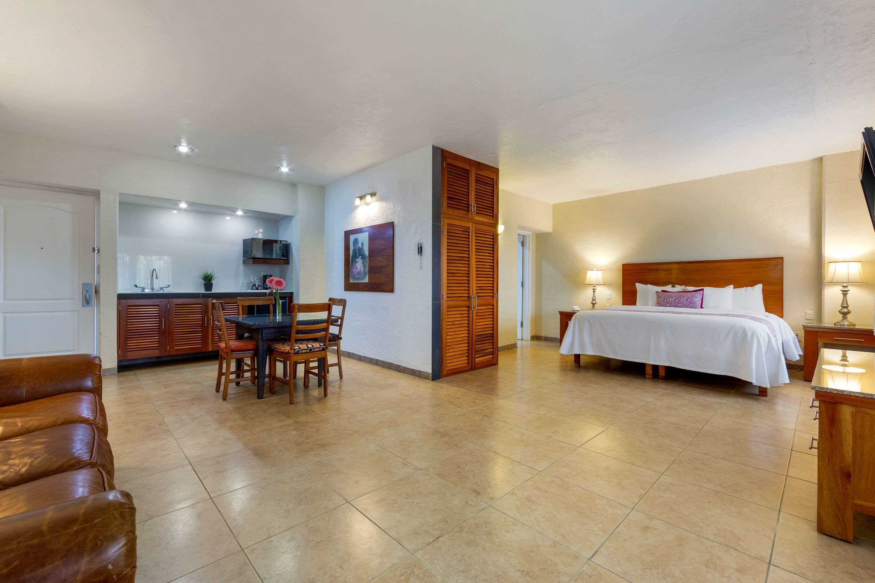 Habitación de hotel con cama, mesita de noche, lámpara y una zona de kitchenette con mesa y sillas.