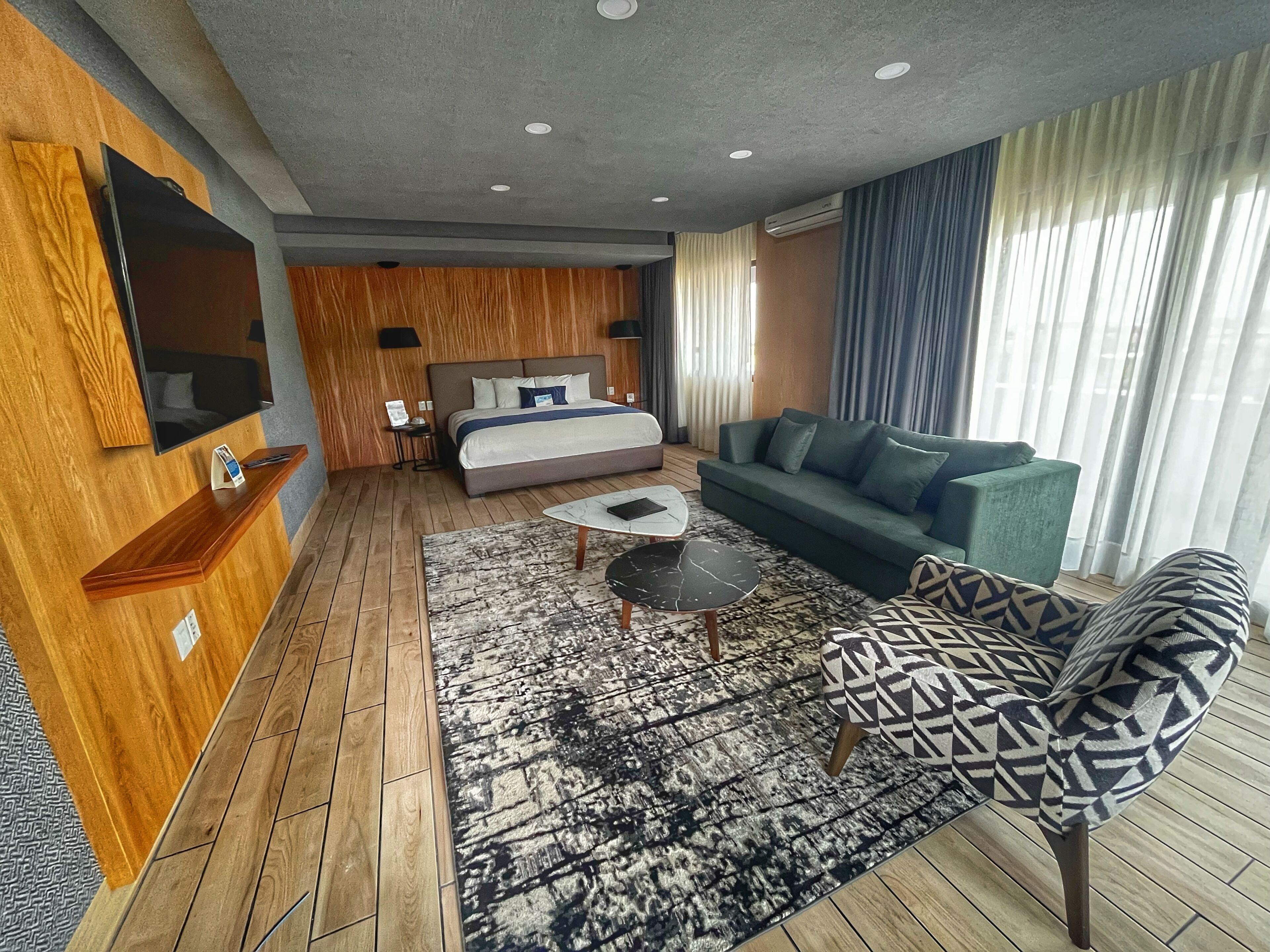 Una habitación de hotel moderna con suelo de madera, una cama grande, un sofá verde, un sillón estampado, una mesa de centro, un televisor de pantalla plana e iluminación empotrada en el techo.