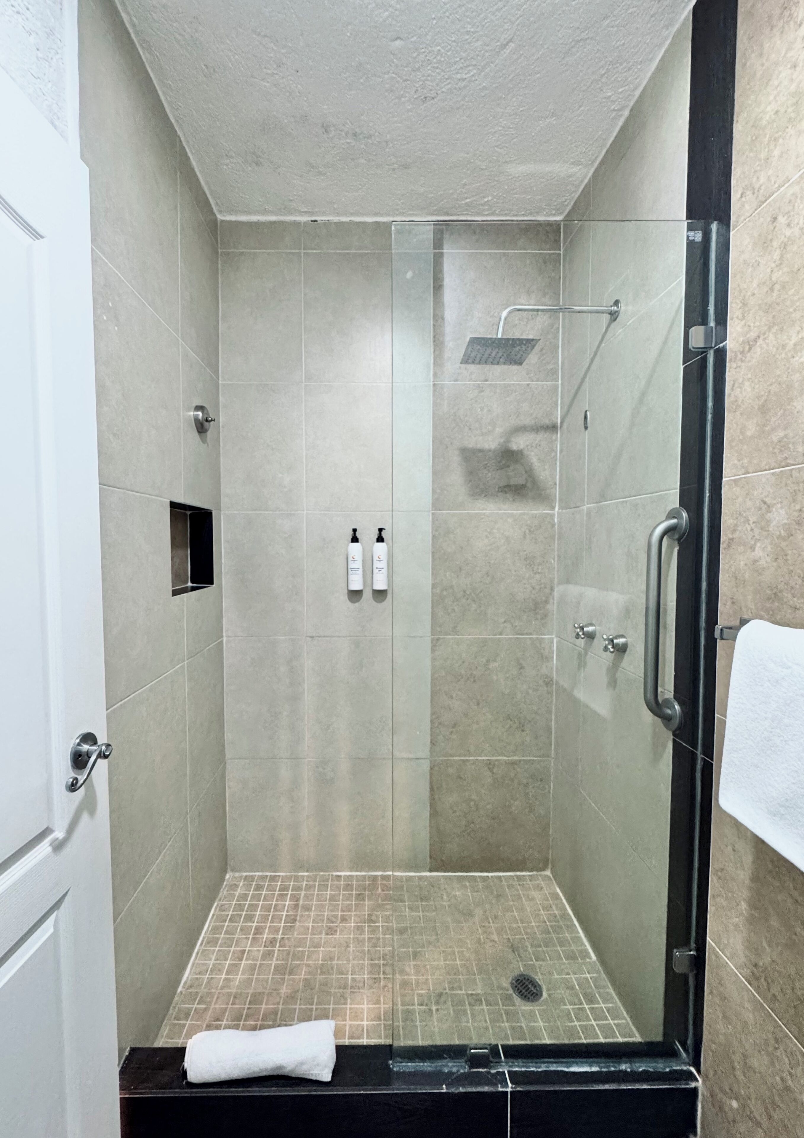 A glass-enclosed shower stall with a tiled floor, a handheld showerhead, and a towel on the floor.