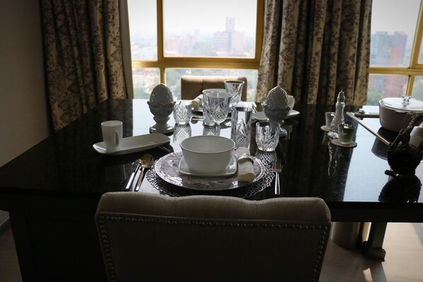 Una mesa de comedor dispuesta con vajilla blanca y copas de cristal, cerca de una ventana con vistas a la ciudad.